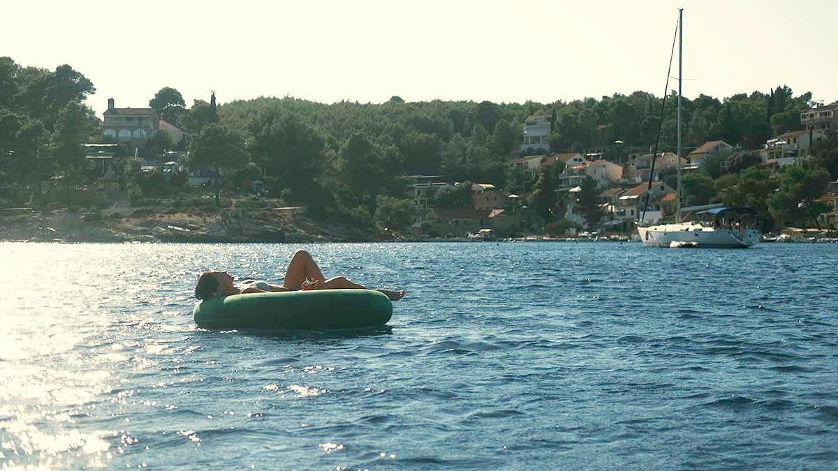 A quiet bay with pine-covered hills, a sailboat, and someone floating on the water